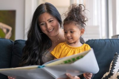 A woman sits on a couch with a small child on her lap, reading a book to her.