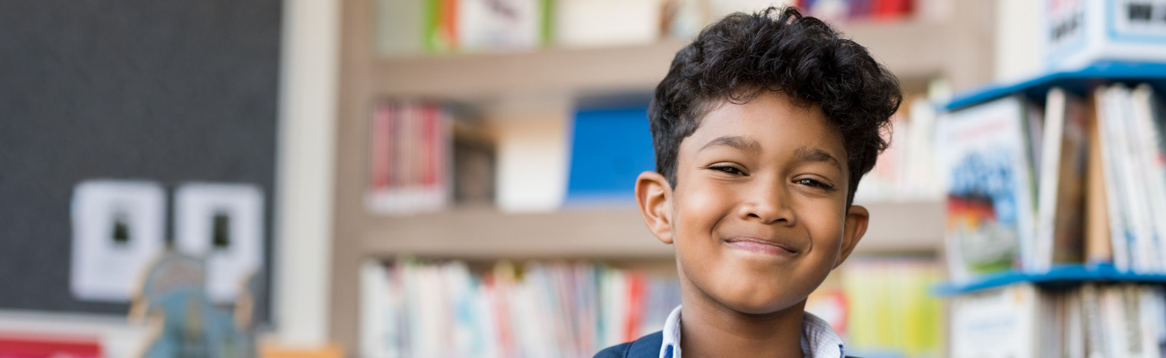 2025 MetaSlider 1300 x 400 Briefs Young bow with brown skin stands in a school library with a backpack on his shoulder and smiles at the camera