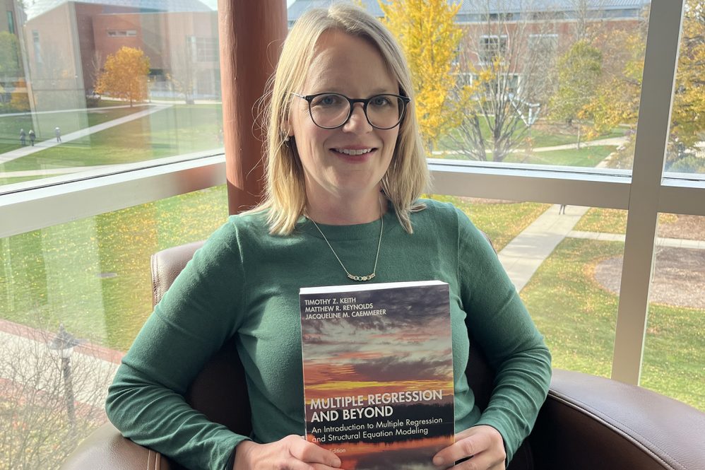 Jacqueline Caemmerer sits in front of a window holding a book titles Multiple regression and Beyond