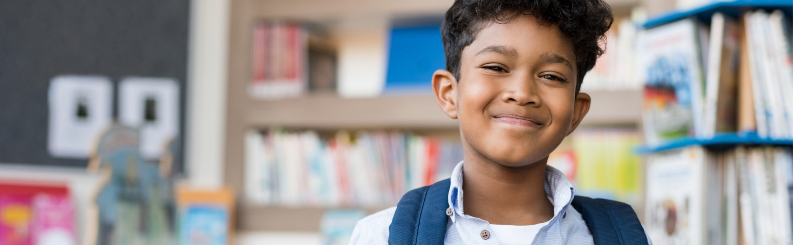 Young boy with brown skin stands in a school library with a backpack on his shoulder and smiles at the camera